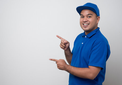 Young smiling asian delivery man in blue uniform and pointing finger on blank space isolated white background.