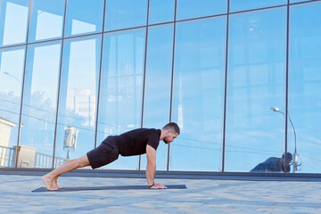 Man athlete in black sportswear practices yoga exercises in plank position outdoors.
