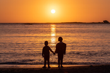 Little girl and little boy  - brother and sister - standing on a Sunset Bay beach against setting sun and holding each other hands, Oregon coast