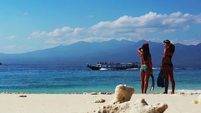 Wide Angle Of Two Girls In Summer Bikinis Talking On The Beach With Seashells On The White Sand As A Small Boat Sails Across The Sea Nearby A Mountain Range, Slowly Tilting Upwards.