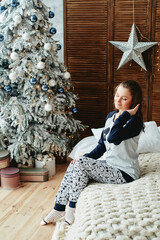 Beautiful woman in pajama sitting on bed in bedroom with Christmas tree and gift boxes on floor, copy space. Cozy home moment. Happy New Year. Christmas concept
