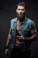 Dressed in modern casual clothing bearded and muscular guy with stylish haircut posing holding beer bottle in dark background.