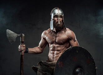 Haired and muscular viking with beard and helmet on him posing holding his axe and shield in dark...