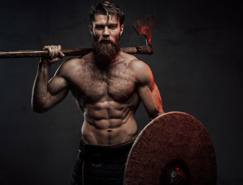 Warlike and furious viking with naked torso and beard staying in dark background holding shield and two handed axe behind his back.