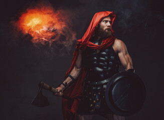 Military bearded roman in red hood and dark armor which holding an axe and shield posing in studio...