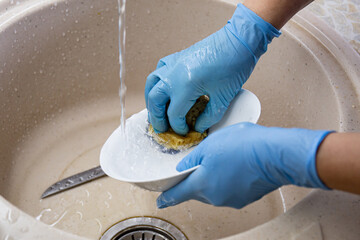 Female hands in blue gloves wash dishes and plates in the kitchen sink. Household concept