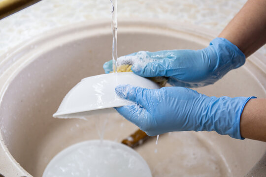 Female Hands In Blue Gloves Wash Dishes And Plates In The Kitchen Sink. Household Concept