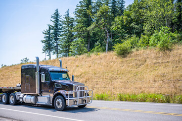 Classic dark big rig semi truck with large grille guard and low cab for better airflow transporting flat bed semi trailer running on the road with hills with trees on the side