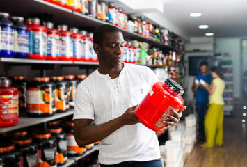 Interested athletically built African buyer choosing for sports nutrition, reading label carefully...