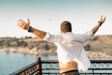 A portrait from the back of a man with his arms raised up against a background of sky and water. The man is wearing a white open shirt