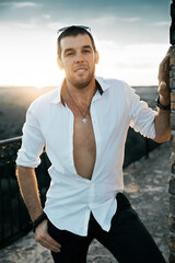 Portrait of 35 year old man in white unbuttoned shirt standing facing camera in warm sunset...