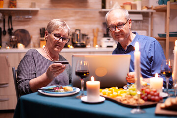 Old woman reading credit card information for online payment while doing shopping with husband. Senior people sitting at the table, browsing, using the technology, internet,