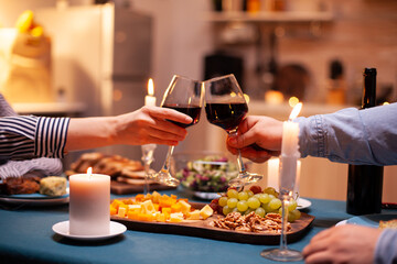 Close up of clinking glasses with wine during romantic dinner dinner celebrating relationship. Happy cheerful young couple dining together in the cozy kitchen, enjoying the meal, celebrating