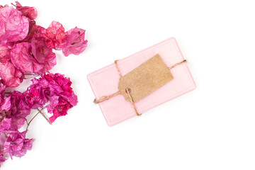 Jabón rosa con etiqueta de papel kraft junto a flores rosa sobre un fondo blanco liso y aislado. Vista superior. Copy space