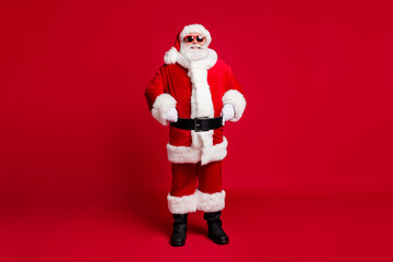 Full length photo of retired old man grey beard self-assured hands hips christmas tree event wear santa costume gloves coat belt sunglass headwear black boots isolated red color background