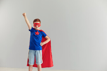 Smiling little boy in a superhero costume raising his hand up on a gray background.