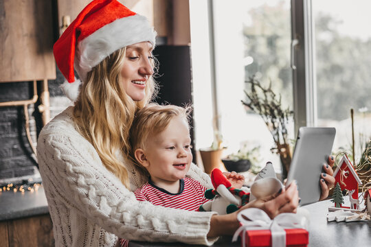 New Year's Video Calls. Mom In A Festive New Year's Santa Hat With A Small Child Sits At A Table With A Tablet And Makes A Video Conference Call. Mother With Baby Looking At The Tablet Screen