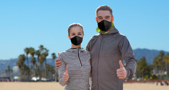 fitness, sport and health concept - couple wearing face protective black reusable masks for protection from virus disease showing thumbs up over venice beach background in california