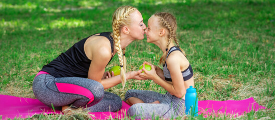 Fototapeta premium Mother kissing her daughter in nose giving green apple sitting on the grass in the park