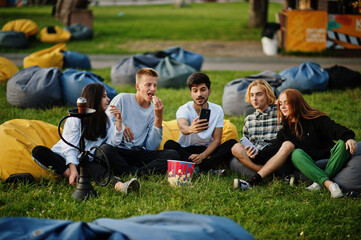 Young multi ethnic group of people watching movie at poof in open air cinema and making selfie on phone.