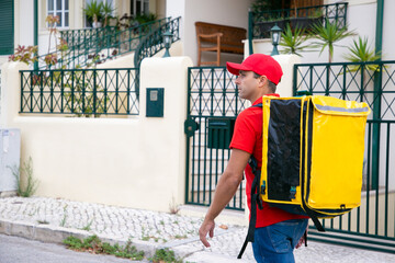 Fototapeta na wymiar Serious deliveryman looking for address and carrying yellow thermal bag. Attractive courier in red shirt walking along street and delivering order. Food delivery service and online shopping concept