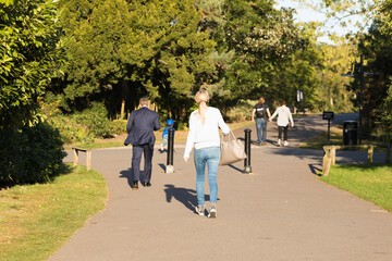 Different Passers-by Walk Along the Asphalt Path of Street Among Green Trees on a Sunny Day. English Park. City People Style, Park Area, Early Autumn. Healthy Walking Every Day. 