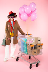 trendy woman in sunglasses and hats with sale tags standing near cart with shopping bags and boxes...