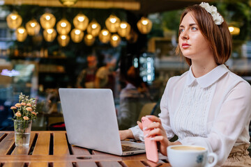 Attractive young elegant caucasian business woman works freelance on laptop computer, sits alone at coffee shop. Looking away. Romantic female blogger. Freelancer thinking about new ideas.