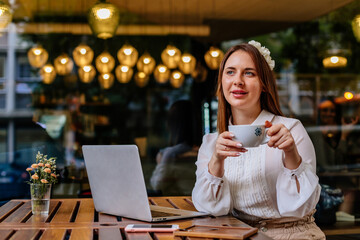 Caucasian romantic woman blogger relaxing drinking coffe while sitting near the window in modern cafe bar, female freelancer thinking about new ideas during work on laptop computer. Looking away 