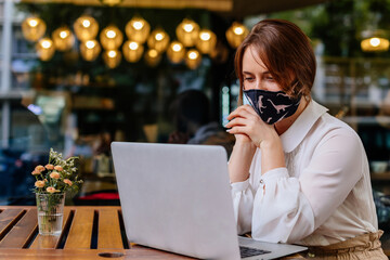 Casual caucasian business woman wearing protective mask working on laptop in a cafe. COVID – 19 virus protection at work. The concept of remote work and online distance learning