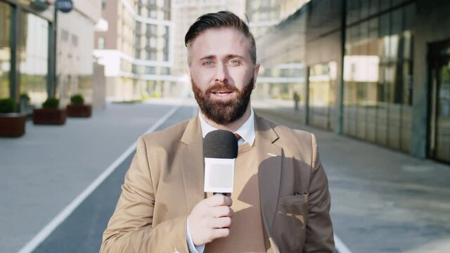 Waist up shot of Caucasian male TV reporter looking at camera and talking into microphone while standing outdoors on city street