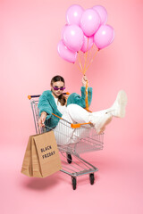 young woman in sunglasses sitting in cart with black friday lettering on shopping bags and holding...