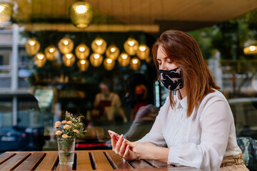 Casual caucasian business woman wearing protective mask using phone for remotely works online in cafe. COVID – 19 virus protection at work. The concept of remote work and online distance learning