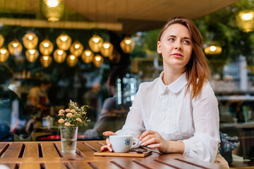 Young beautiful woman holding a cup of coffee in cafe. Outdoor portrait of female blogger posing while traveling holiday vacation. Happy freelancer thinking about new ideas.