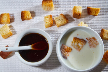 Square toasted pieces of homemade delicious rusk, hardtack, Dryasdust, zwieback, Liquid honey in a plate and cap of milk on a white tablecloth.
