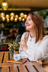 Young beautiful woman holding a cup of coffee in cafe. Outdoor portrait of female blogger posing while traveling holiday vacation. Happy freelancer thinking about new ideas.