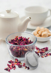 Fresh organic rose petals with strainer infuser and white ceramic teapot and cane sugar on white.
