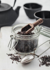Black loose organic tea in glass jar with scoop and strainer infusor on light background with iron teapot and cups.