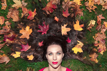 Portrait of a brunette woman with long hair. She lies on green grass in a red dress and maple leaves in her hair. Autumn concept ..