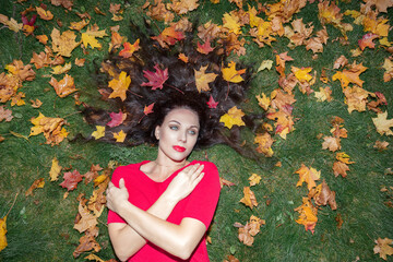 Portrait of a brunette woman with long hair. She lies on green grass in a red dress and maple leaves in her hair. Autumn concept ..