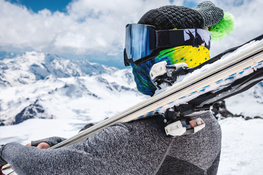 Close-up young woman skier in a ski mask with a closed face holds skis on her shoulder against the backdrop of snow-capped mountains