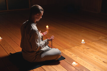 An attractive caucasian man in a lotus position sits in a dark meditation room and holds a red rosary for prayer in his hand against the background of burning candles