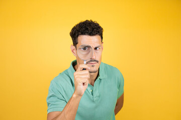 Young handsome man wearing green casual t-shirt over isolated yellow background surprised looking through a magnifying glass