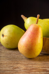 Close-up of focused ercolina pear and several in defocused wooden bowl, on wooden table and black background in vertical, with copy space