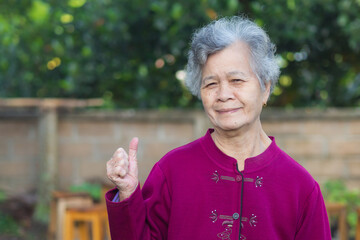 Senior Asian woman with short white hair smiling and showing thumbs up while standing in a garden. Concept of old people and healthcare
