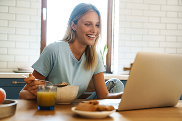 Obraz na płótnie Canvas Beautiful joyful girl using laptop and eating cereal