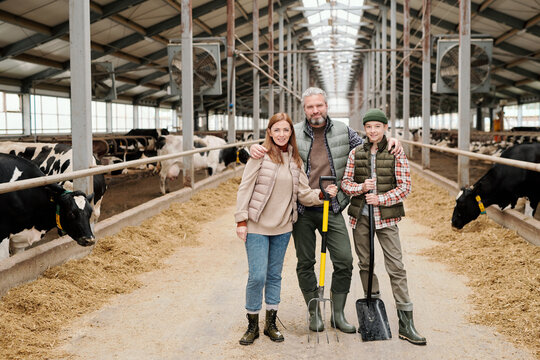 Portrait of content contemporary family of farmers standing with work tools in large cowshed
