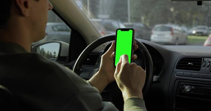 Over the shoulder view. Handsome man in a sweatshirt sitting in the car and using iPhone 11 with green screen, tapping, scrolling, swiping. Work from a vehicle. Fall background.
