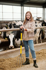 Portrait of smiling attractive female farmhouse owner in vest standing against cows in stall and working with pitchfork © pressmaster