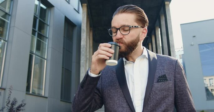 Front view of positive good-looking successful smiling adult businessman with well-groomed beard in glasses which drinking coffee near modern urban office building